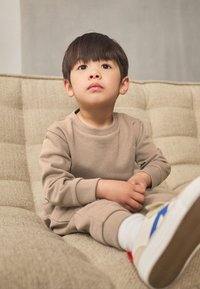 Set de trening din bumbac bej, format din bluza și pantaloni, purtat de un copil așezat pe o canapea texturată de culoare deschisă. Pantofii au accente colorate și talpă albă.