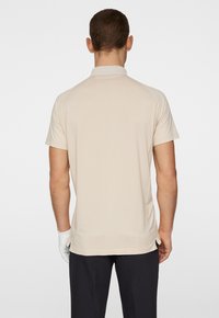 Beige kortärmad polo med slät yta, krage, rak fåll och detaljerad bakpanel. Bärs med svarta byxor och en vit handske.