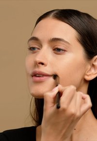 En makeupbørste i en hånd påfører produkt på en kind. Personen har glat hud og mørkebrunt hår, med blødt lys og en neutral baggrund.