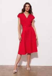 Czerwona sukienka z dekoltem w serek, krótkimi rękawami i dopasowanym pasem z dwoma guzikami. Rozkloszowana spódnica o gładkiej teksturze; modelka nosi srebrne szpilki.