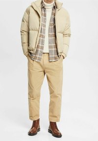 Beige Steppjacke, ein kariertes Hemd in Grau und Braun, cremefarbener Pullover und hellbraune Hose. Braune Schnürstiefel vervollständigen das Outfit.