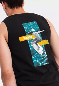 Svart ärmlös tröja med ett grafikmotiv av en surfare på en surfbräda över ett mönster av blått vatten, med gult "Sommar"-text och detaljer.