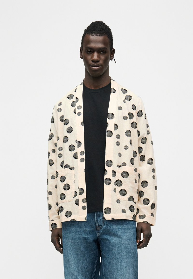 Άνδρας φορώντας ανοιχτό μπεζ σακάκι με μεγάλα μαύρα κυκλικά σχέδια, μαύρο πουκάμισο και μπλε τζιν μπροστά σε απλό φόντο.