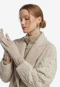 Beiga stickade handskar med en mjuk textur, med en etikett på handleden. Bärs av en person, ihop med en ljusfärgad ullrock och tröja.