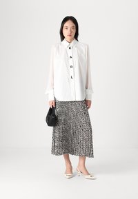 Chemise blanche à manches transparentes avec le lettrage "KARL", associée à une jupe plissée noire et blanche à motifs et un sac à main noir.