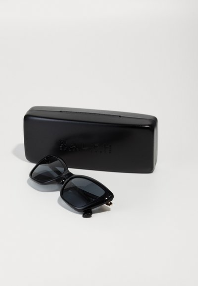 Gafas de sol negras de gran tamaño con lentes oscuros, forma rectangular y montura de plástico brillante, al lado de un estuche rígido negro con texto en relieve.
