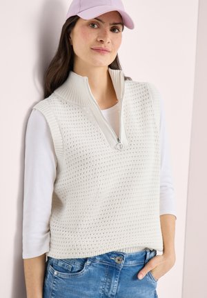 Frau mit einer lavendelfarbenen Mütze, weißer Strickweste mit Reißverschlusskragen über einem weißen Langarmshirt und blauen Jeans, stehend mit einer Hand in der Tasche.