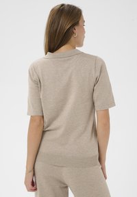 Pull polo beige à manches courtes avec ourlet et col côtelés. Texture douce, coupe décontractée et détails de couture visibles sur les manches et l'ourlet.