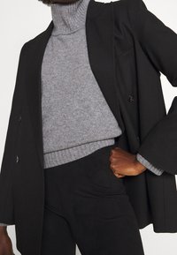 Zwarte dubbelzijdige blazer over een grijze coltrui, met ribbeltextuur en aangesloten ontwerp, aangevuld met zwarte broek.