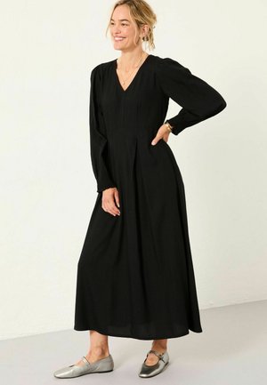 Schwarzes langärmliges Kleid mit V-Ausschnitt, gerafften Ärmeln und plissiertem Rock. Kombiniert mit silbernen flachen Schuhen auf hellem Hintergrund.