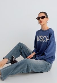 Μπλε φούτερ με το λογότυπο "MSCH", άνετη εφαρμογή· φαρδιά τζιν παντελόνια σε ανοιχτό μπλε χρώμα· σκούρα γυαλιά ηλίου· ομαλή υφή· λιτή σχεδίαση.