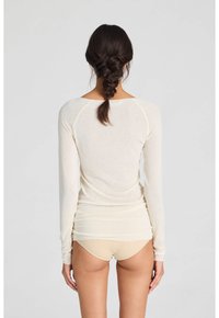 Cămașă crem cu mâneci lungi din material ușor, cu un design ajustat. Are mâneci raglan și un tiv curbat, purtată cu pantaloni bej.