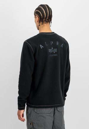 Persoon met gevlochten haar die een zwart sweatshirt draagt met de tekst en het logo "ALPHA INDUSTRIES" op de rug, gecombineerd met grijze cargobroek.