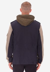 Uomo visto di spalle che indossa una giacca blu navy con maniche beige e cappuccio marrone attaccato, in piedi davanti a uno sfondo chiaro e uniforme.