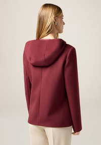 Giacca con cappuccio bordeaux dal design strutturato, texture liscia e maniche lunghe, caratterizzata da una cucitura centrale sul retro e dettagli minimi.