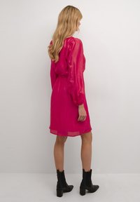 Rochie în nuanță fuchsia, cu manșoane lungi și pufoase și talie strânsă, confecționată din material transparent. Poartă cizme negre până la gleznă, cu toc gros.