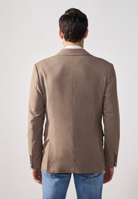 Brown tailored blazer with a smooth texture, single vent at the back, and metallic buttons on the sleeves. Worn with light blue jeans.