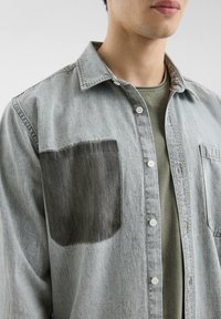Hellgrau Denim-Hemd mit einem auffälligen schwarzen Patch auf der linken Brusttasche, weißen Knöpfen und einem Kragen.