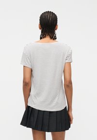 Άτομο με πλεξούδες που φορά λευκό πουκάμισο κοντών μανικιών με λεπτές μαύρες ρίγες και μαύρη πλισέ φούστα, εμφανιζόμενο από πίσω.