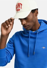 Blauwe hoodie met een trekkoord, met een logo-badge op de borst. Crème kleurige pet met rode tekstdetails. Close-up van handen die de pet aanpassen.