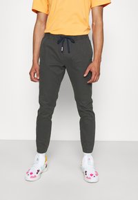 Donkergrijze joggingbroek met een trekkoord in de taille, gecombineerd met witte sneakers met kleurrijke ontwerpen. Het shirt is oranje.