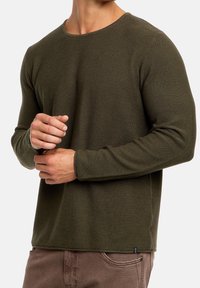 Suéter texturizado de manga larga en verde oliva con cuello redondo. Presenta puños y dobladillo acanalados, combinado con pantalones marrones. Etiqueta de la marca sutilmente visible.