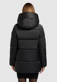 Geacă neagră tip puffer cu glugă, având secțiuni largi matlasate, material exterior moale și un logo pe spate.