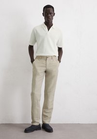 Polo shirt in light cream with short sleeves, paired with beige linen trousers and dark loafers. Model stands in front of a neutral background.