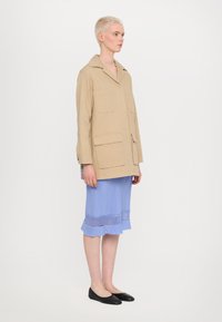 Beige oversized kabát se dvěma předními kapsami, kombinovaný s světle modrou sukní s volánkovým lemem a síťovaným akcentem. Ploché černé boty.