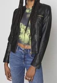 Czarna skórzana kurtka o dopasowanym kroju, z prążkowanymi akcentami na ramionach i złotymi detalami zamka błyskawicznego, zestawiona z kolorowym, krótkim topem i niebieskimi dżinsami.