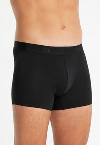 Boxer-briefs noirs en tissu lisse. Dotés d'une large ceinture noire avec un texte de marque. L'avant présente un design de poche contournée.