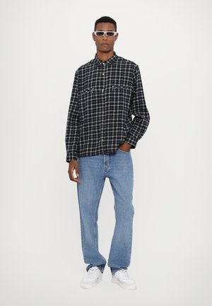 Camicia in flanella a scacchi bianco e nero con maniche lunghe, bottoni davanti, due tasche sul petto, abbinata a jeans azzurri chiari e sneakers bianche.