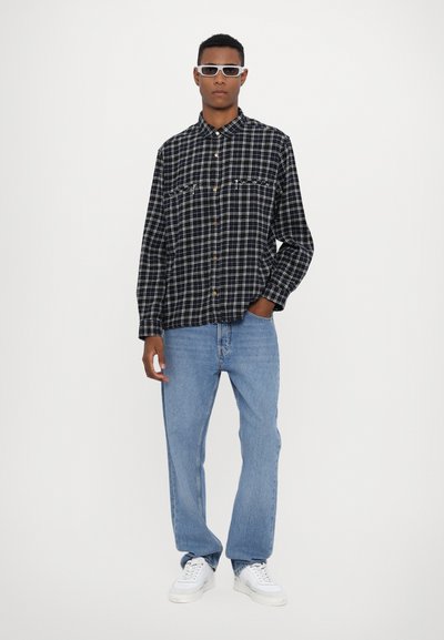 Camicia in flanella a scacchi bianco e nero con maniche lunghe, bottoni davanti, due tasche sul petto, abbinata a jeans azzurri chiari e sneakers bianche.
