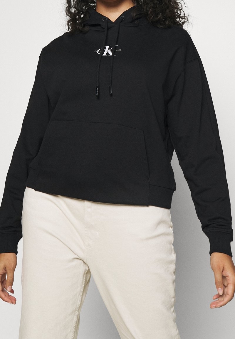 Sudadera negra con capucha que presenta un bolsillo frontal, puños elásticos, un diseño corto y un pequeño logo en el pecho. Fabricada con tejido suave.