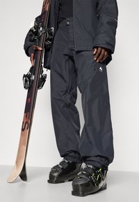 Schwarze Skihosen aus wasserabweisendem Material mit elastischen Bündchen. Kombiniert mit schwarzen Skischuhen mit grünen Akzenten und einem Ski mit Bindungen.