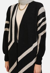Long black and cream cardigan with a geometric striped pattern, ribbed cuffs, and a deep V-neck design. Made from textured fabric.