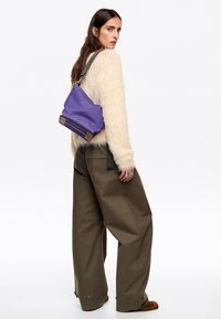 Fioletowa torba o luźnym fasonie, z brązowymi akcentami na dnie i szarym regulowanym paskiem, noszona na ramieniu na jasnym tle.