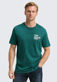 Camiseta de algodón corta de manga azul en tonos verde azulado con cuello redondo. Presenta texto y gráfico en blanco en el pecho izquierdo. Combinada con jeans oscuros.