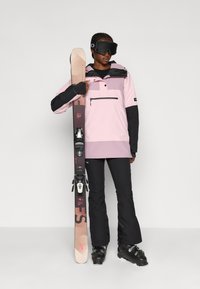 Suusatamisriietus sisaldab roosa ja musta jopega, millel on ees tasku, musti pükse ja lumiseid saapaid; isik hoiab suuski kinnitustega.