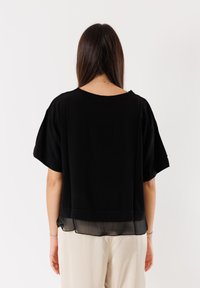 Maglietta nera con maniche corte, realizzata in un tessuto morbido e con un orlo trasparente. Indossata con pantaloni chiari, che mostrano una vestibilità rilassata e un design semplice.
