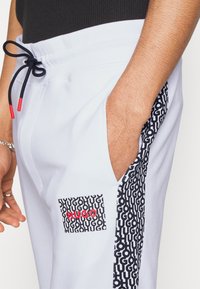 Weiße Jogginghosen mit einem schwarz-weißen geometrischen Muster an der Seite, einem roten Logo-Patch und einem schwarzen Kordelzug in der Taille.