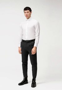 Camicia bianca con bottoni, pantaloni neri su misura, cintura nera e stivaletti. Tessuto liscio con leggero lucido, design aderente e aspetto ordinato.