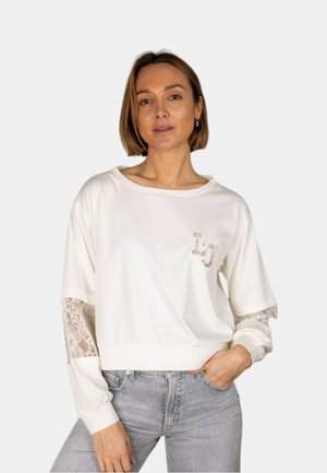 Donna che indossa una felpa bianca con inserti in pizzo sulle maniche e le iniziali "LJ", abbinata a jeans grigio chiaro, mano in tasca.