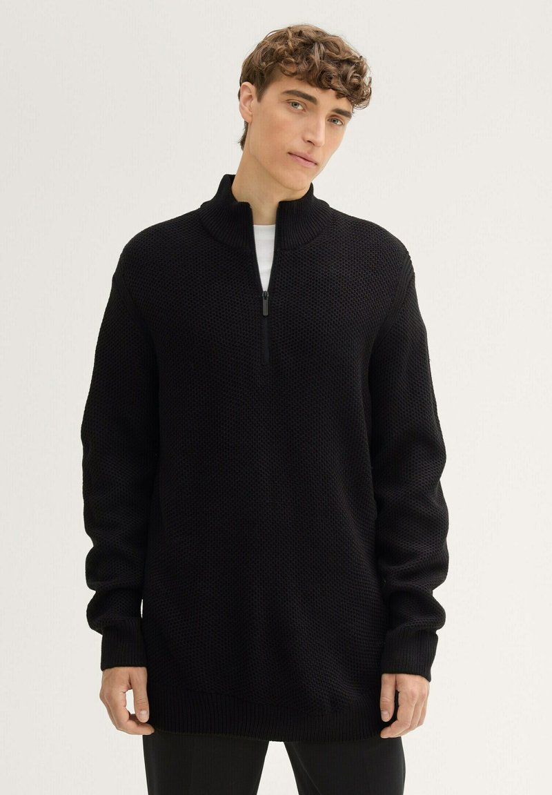 Schwarzer Strickpullover mit einem halben Reißverschluss am Kragen, der ein strukturiertes Muster sowie gerippte Bündchen und Saum aufweist. Lässiger, lockerer Schnitt.