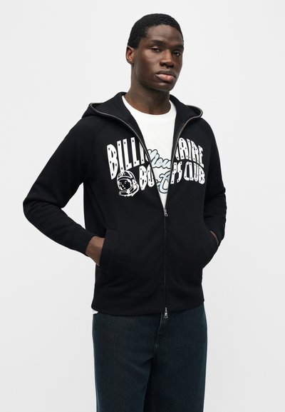 Νεαρός άνδρας που φοράει μαύρο φούτερ με φερμουάρ και το κείμενο "Billionaire Boys Club" μαζί με το λογότυπο αστροναύτη, λευκό τυπωμένο μπλουζάκι και σκούρο τζιν, με τα χέρια στις τσέπες.