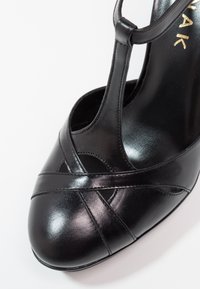 Chaussure à talon haut en cuir noir avec un design à T, détails entrecroisés sur le bout et texture lisse et brillante.
