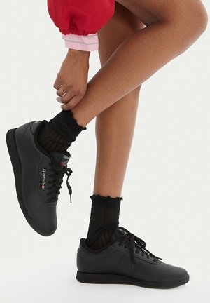 Osoba nosząca czarne trampki Reebok i przezroczyste czarne skarpetki, poprawiająca jedną stopę, z widoczną długą czerwoną i różową rękawicą.