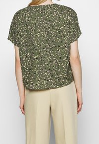 Blusa verde a maniche corte con un motivo floreale in viola, nero e crema. Taglio morbido con orlo arrotondato, abbinata a pantaloni beige.