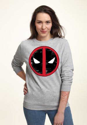 Kobieta o długich brązowych włosach nosi szarą bluzę z czerwono-czarnym logo Deadpool oraz niebieskie dżinsy, stoi z ręką na biodrze.