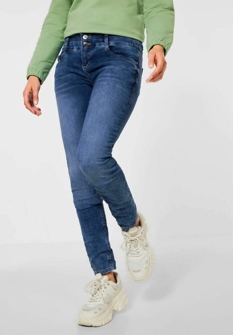 Mörkblå denimjeans med slim fit, med två framfickor och knappstängning. Bärs med sneakers i krämfärg.